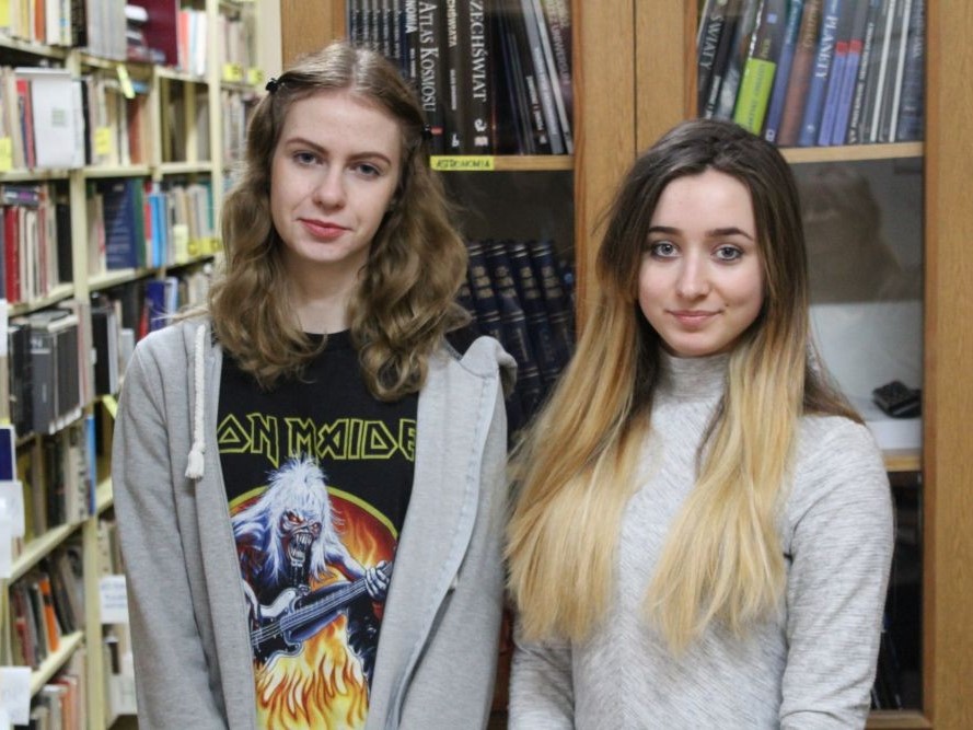 Dwie uczennice pozują na tle książek w bibliotece