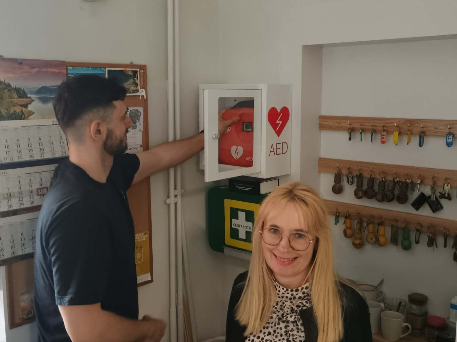 Mężczyzna i kobieta stoją obok siebie. Mężczyzna wkłada pomarańczowy defibrylator w opakowaniu do białej skrzynki. Kobieta się uśmiecha