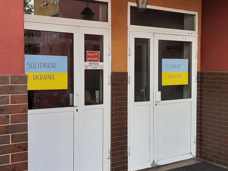 Widoczne  szklane drzwi do szkoły z plakatem w formie flagi Ukrainy. Na plakacie napis: Solidarni z Ukrainą