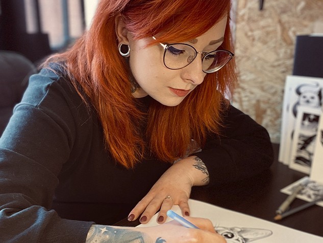 Kobieta w okularach, w długich włosach rysuje coś przy stole. Na stole znajdują się rysunki zwierząt 