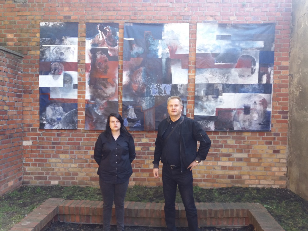 Mężczyzna po prawej stronie, po lewej kobieta. Oboje stoją. Za nimi ściana z pracami w formie obrazów.