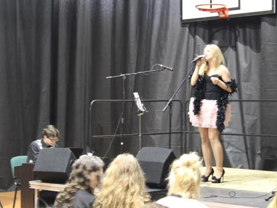 Artystka na scenie śpiewa do mikrofonu, akompaniuje chłopak przy fortepianie, na widowni widać trzy osoby