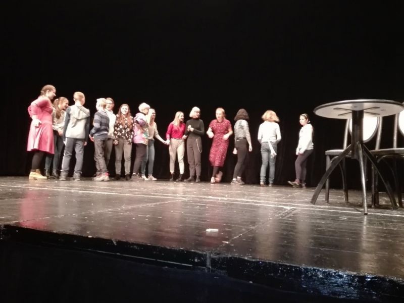 Grupa młodzieży, kilkanaście osób stoi w jednym szeregu, lekkim półkolem. Wśród nich aktor coś pokazuje. Całość jest na scenie teatru.
