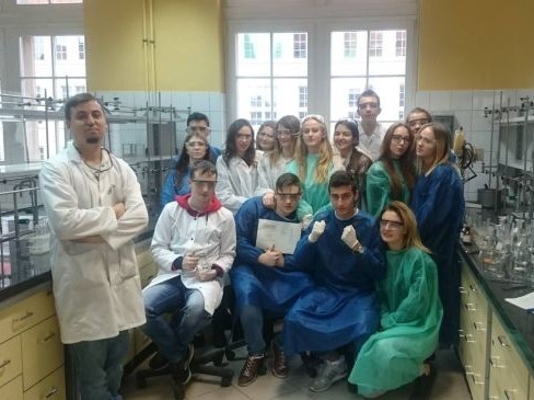 Grupa kilkunastu uczennic i uczniów stoi pomiędzy stołami laboratoryjnymi i pozuje do wspólnego zjęcia. Wszyscy są ubrani w jednorazowe fartuchy