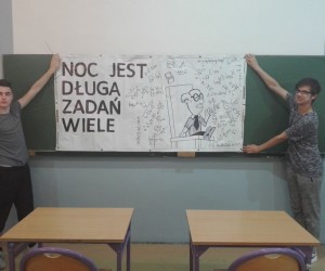 Dwóch uczniów stoi po dwóch stronach banera, trzymają go za końce. Na banerze widać napis:
Noc jest długa, zadań wiele.