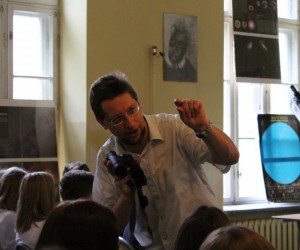 Wykladowca dr Tomasz Stechnij pochyla się nad siedzącymi uczniami. W prawej ręce trzyma lornetkę, lewą ręką, dwoma palcami pokazuje wielkość jakiegoś obiektu.