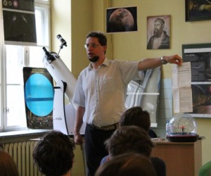 Wykładowca, dr Tomasz Stechnij lewą ręką coś wskazują w bok od siebie. Stoi na tle okna, zdjęć astronomicznych. Przed nim widać głowy uczniów - trzech.