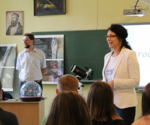 Po prawej stronie stoi kobieta, która mówi i uśmiecha się. Po lewej stronie stoi wykładaowca i się uśmiecha. Przed nimi, na pierwszym planie widać kilka głów młodych ludzi. Młodzi ludzie siedzą.