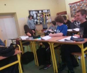 Dwóch uczniów stoi pod ścianą. Jeden ma głowie ławkę. Uczniowie ci coś pokazują.. Kilku uczniów siedzi bokiem, obrócona twarzami do demonstrujących.