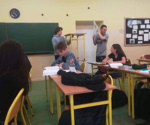 Widać jednego ucznia trzymającego w górze ekierkę. Obok niego stoją dwie ławki jedna na drugiej. Za ławkami stoi drugie uczeń. Widać trójkę uczniów siedzących w ławkach.