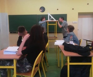 Dwóch uczniów na tle zielonej tablicy. Pomiędzy nimi są dwie ławki ustawione jedna na drugiej. Do ławki górnej i tablicy przyłożona jest duża, szkolna ekierka. Kilkoro uczniów siedzi w ławkach i patrzy na tych pod tablicą.